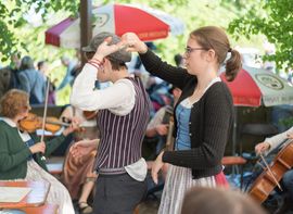 Ein junger Mann und eine junge Frau in Dirndl und Weste tanzen unter Sonnenschirmen im Freien zu Live-Volksmusik